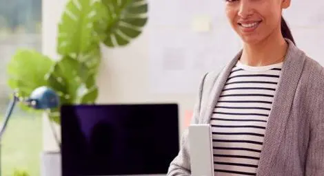 A woman is smiling at the camera and standing in front of a desk with a computer on it.