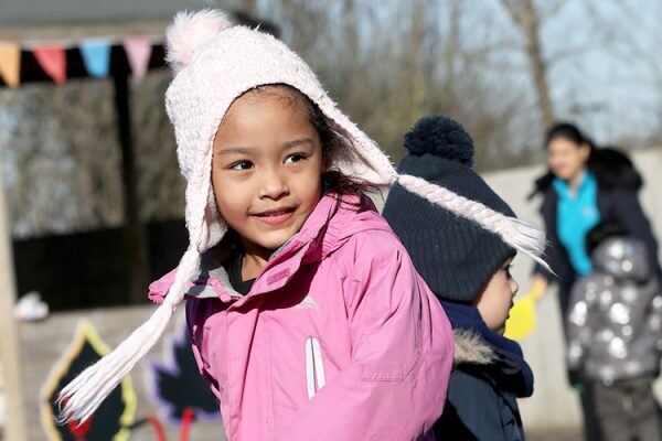 A young girl wearing a pink jacket and wooly hat is walking through a playground.