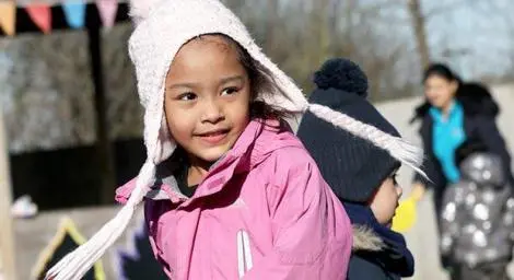 A young girl wearing a pink jacket and wooly hat is walking through a playground.